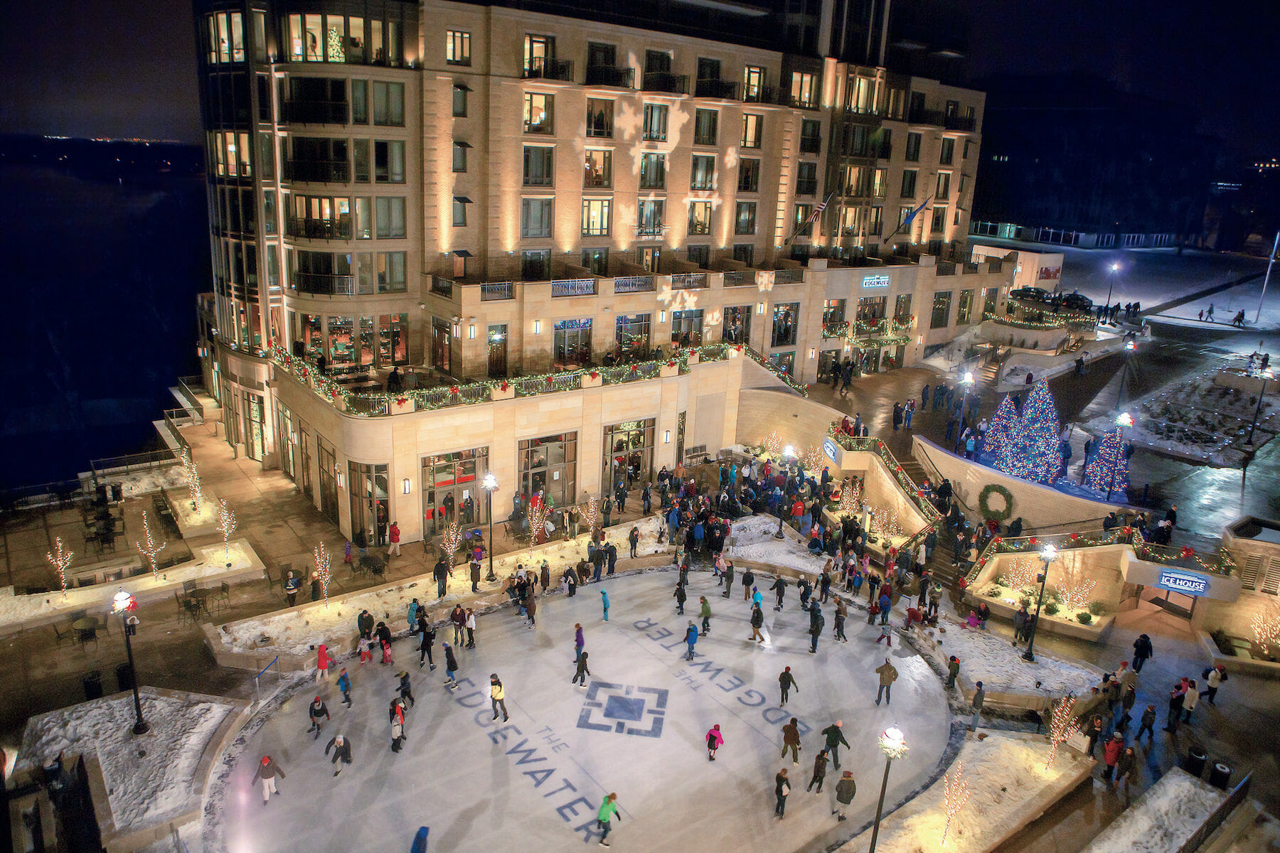 Skaters enjoy a lively ice rink surrounded by festive lights and decorated trees at a bustling waterfront location.