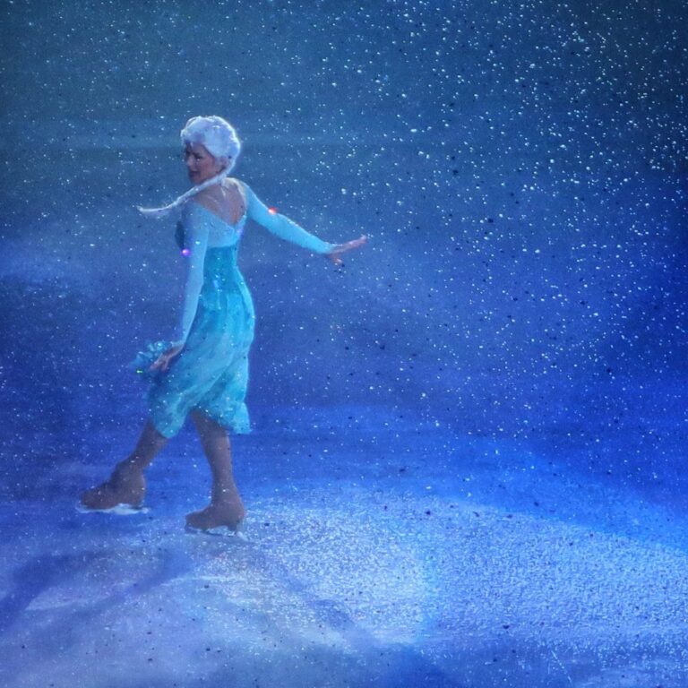 A figure skater in a shimmering blue dress glides gracefully on ice, surrounded by swirling snowflakes and blue light.